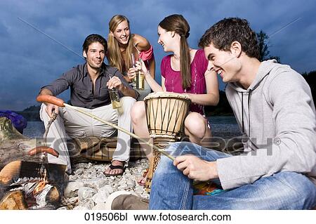 Stock Image - group of friends enjoying beer and sausages at party on riverbank in summer. Fotosearch