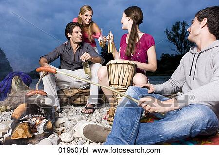 Stock Image - group of friends enjoying beer and sausages at party on riverbank in summer. Fotosearch