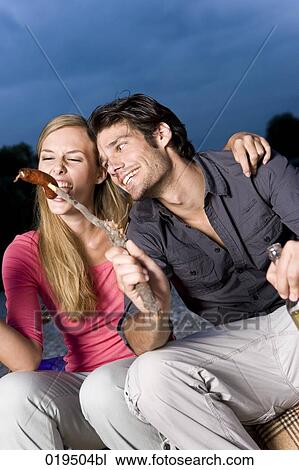 Stock Image - portrait of young couple sharing sausage at party on riverbank in summer. Fotosearch