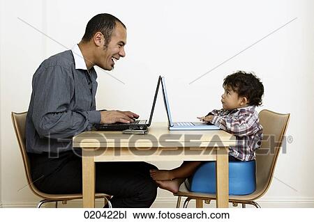 Stock Image - man working from home sitting at table with little son. Fotosearch