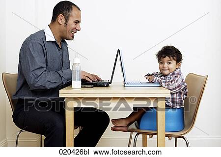 Stock Image - man working from home sitting at table with little son. Fotosearch
