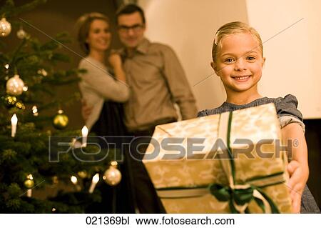 portrait of young girl holding christmas present with parents in background View Large Photo Image Stock Image - portrait of young girl holding christmas present with parents in background . Fotosearch
