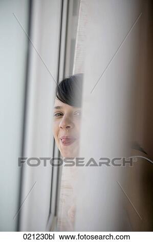 Stock Image - portrait of young woman standing at window sticking out tongue. Fotosearch