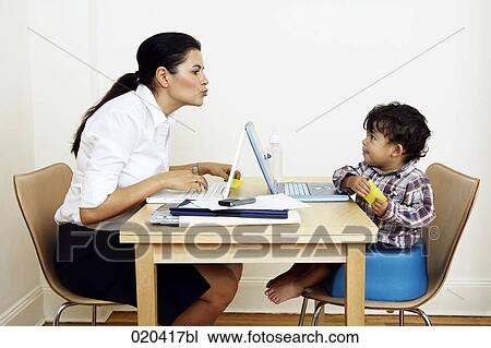 Stock Image - woman working from home sitting at table with little son. Fotosearch