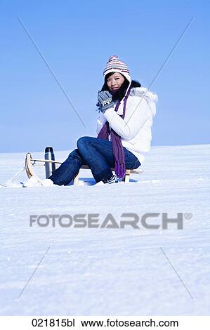 Stock Image - young asian woman sitting on sledge having drink from thermos bottle. Fotosearch