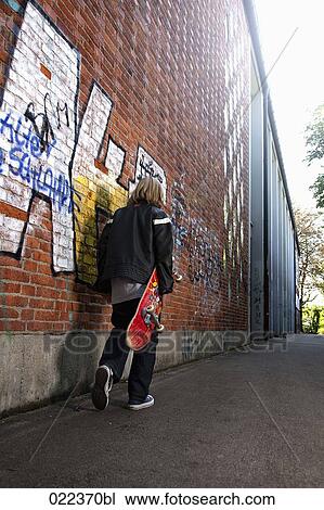 Stock Image - rear view of young boy walking along wall with skatebord. Fotosearch