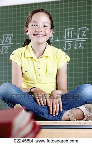 Stock Image - young girl sitting on desk in front of blackboard . Fotosearch