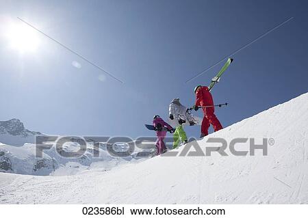 Stock Image - group of three people with skis and snowboards walking up mountain. Fotosearch