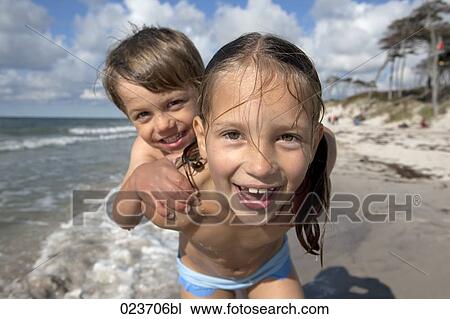 Stock Image - portrait of young girl giving piggyback ride to little brother on the beach. Fotosearch