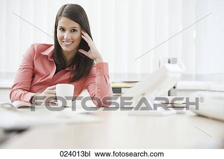 Stock Image - portrait of young businesswoman sitting at desk with cup. Fotosearch
