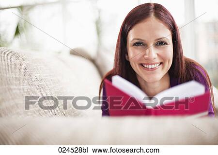 Stock Image - portrait of young woman lying on sofa reading book. Fotosearch