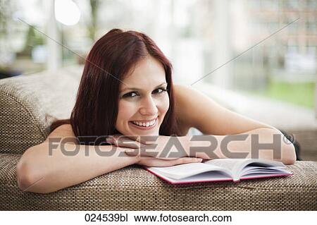 Stock Image - portrait of young woman lying on sofa with book. Fotosearch