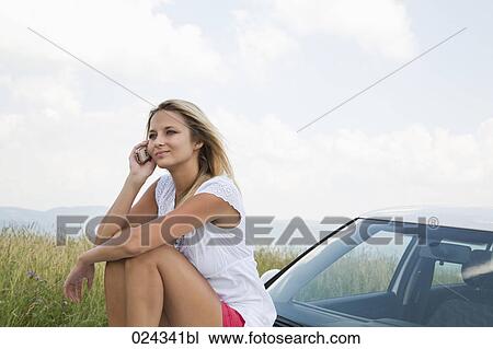 Stock Image - portrait of young woman siiting on bonnet of car talking on mobile phone. Fotosearch