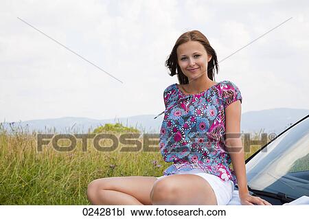 Stock Image - portrait of young woman sitting on bonnet of car. Fotosearch