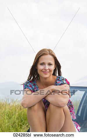 Stock Image - portrait of young woman sitting on bonnet of car. Fotosearch