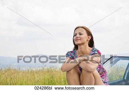 Stock Image - portrait of young woman sitting on bonnet of car. Fotosearch