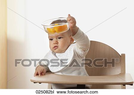 Stock Image - portrait of baby in high chair holding bowl with fruit. Fotosearch