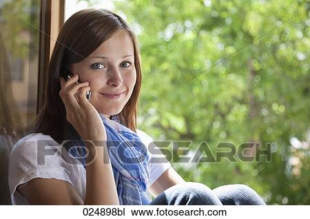 Stock Image - portrait of young woman talking on mobile phone at window. Fotosearch