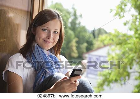 Stock Image - portrait of young woman with mobile phone at window. Fotosearch