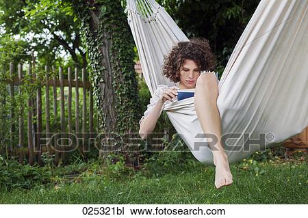 Stock Image - young woman lying in hammock in garden reading. Fotosearch