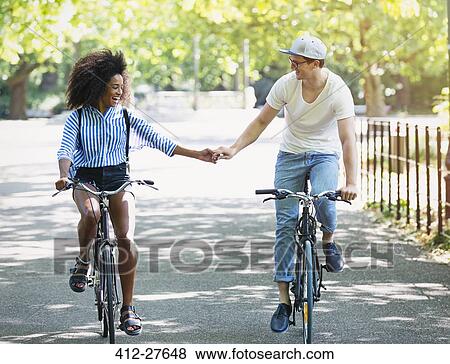 Stock Photo - Couple holding hands riding bicycles in urban park. Fotosearch