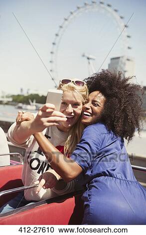 Stock Image - Enthusiastic friends taking selfie on double-decker bus with London Eye in background. Fotosearch