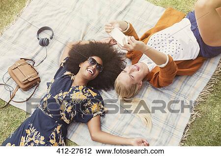 Stock Image - Friends taking selfie relaxing on blanket in grass. Fotosearch