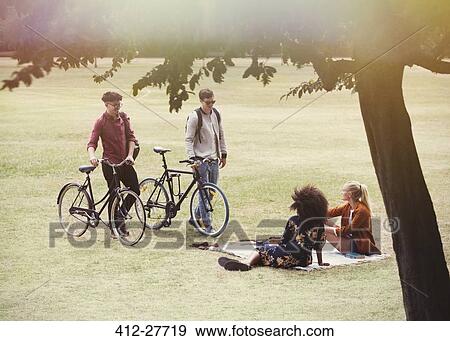 Stock Photo - Men with bicycles approaching women on blanket in park. Fotosearch