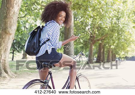 Stock Image - Portrait smiling woman riding bicycle with mp3 player and headphones in park. Fotosearch