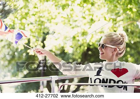 Stock Image - Smiling woman waving British flag on double-decker bus. Fotosearch