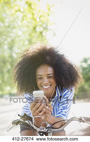 Picture - Smiling woman with afro listening to music with headphones and mp3 player on bicycle. Fotosearch