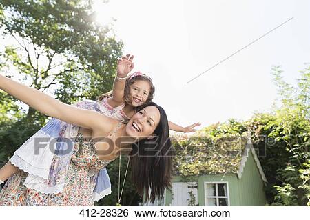 Stock Photograph - Carefree mother piggybacking daughter with arms outstretched in backyard. Fotosearch