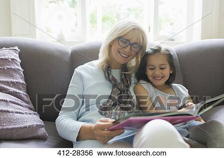 Stock Photograph - Grandmother and granddaughter reading book on living room sofa. Fotosearch
