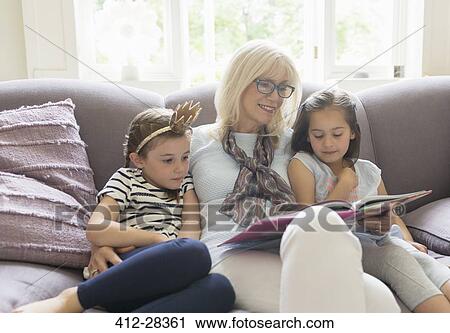 Stock Image - Grandmother and granddaughters reading book on living room sofa. Fotosearch