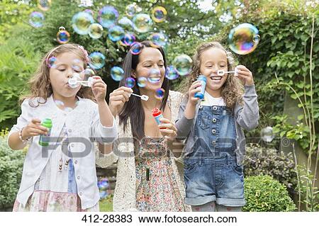 Stock Image - Mother and daughters blowing bubbles in backyard. Fotosearch