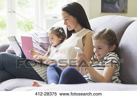 Stock Photography - Mother and daughters with laptop, cell phone and digital tablet on sofa. Fotosearch