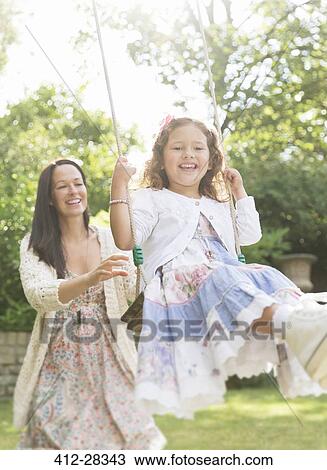 Stock Image - Mother in dress pushing daughter on swing in backyard. Fotosearch