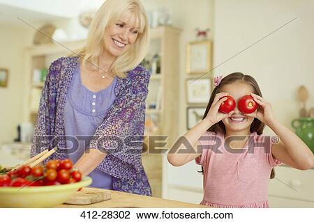 Stock Image - Portrait playful granddaughter covering eyes with tomatoes in kitchen. Fotosearch