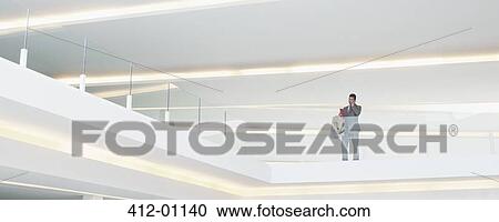 Stock Image - Businessman at glass balcony railing in modern office. Fotosearch
