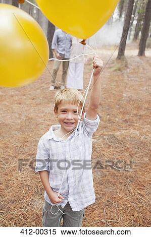 Portrait of smiling boy holding balloons in woods with parents in background View Large Photo Image Stock Photography - Portrait of smiling boy holding balloons in woods with parents in background. Fotosearch
