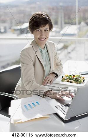 Stock Photography - Portrait of smiling businesswoman eating lunch and working at desk. Fotosearch