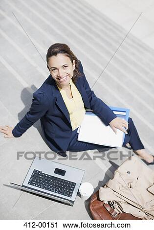Stock Image - Portrait of smiling businesswoman with paperwork, coffee and laptop on steps. Fotosearch