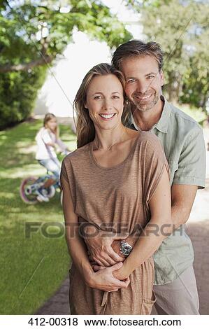 Portrait of smiling couple hugging in driveway with daughter on bicycle in background View Large Photo Image Stock Photo - Portrait of smiling couple hugging in driveway with daughter on bicycle in background. Fotosearch