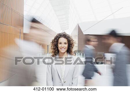 Portrait of smiling woman with co-workers rushing by in lobby View Large Photo Image Stock Image - Portrait of smiling woman with co-workers rushing by in lobby. Fotosearch