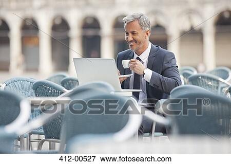 Stock Photo - Smiling businessman drinking coffee and using laptop at sidewalk cafe. Fotosearch