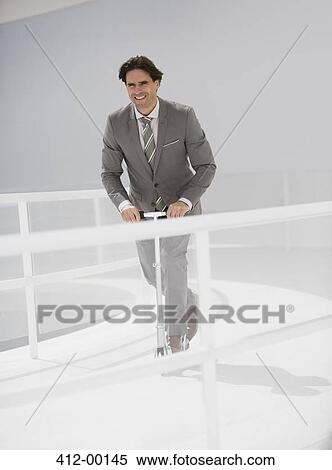Stock Photography - Smiling businessman riding scooter along walkway. Fotosearch