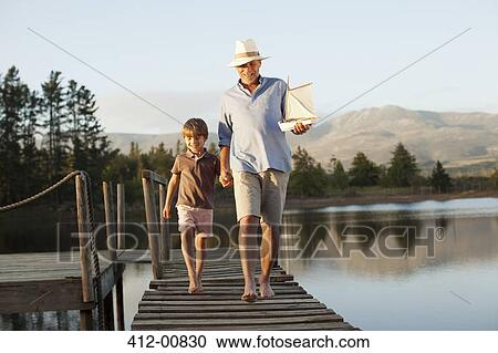 Stock Image - Smiling grandfather and grandson with toy sailboat holding hands and walking along dock over lake. Fotosearch