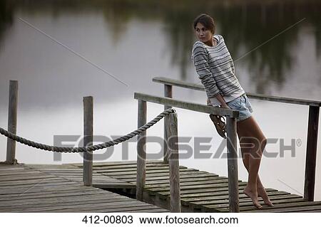 Stock Image - Woman leaning on railing on dock over lake. Fotosearch