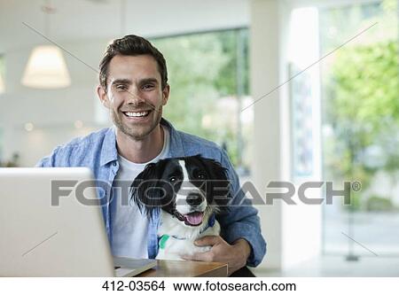 Picture - Man with dog using laptop in kitchen. Fotosearch
