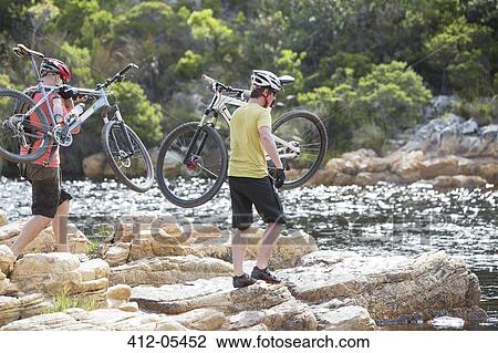 Stock Image - Men carrying mountain bikes on rock formation. Fotosearch